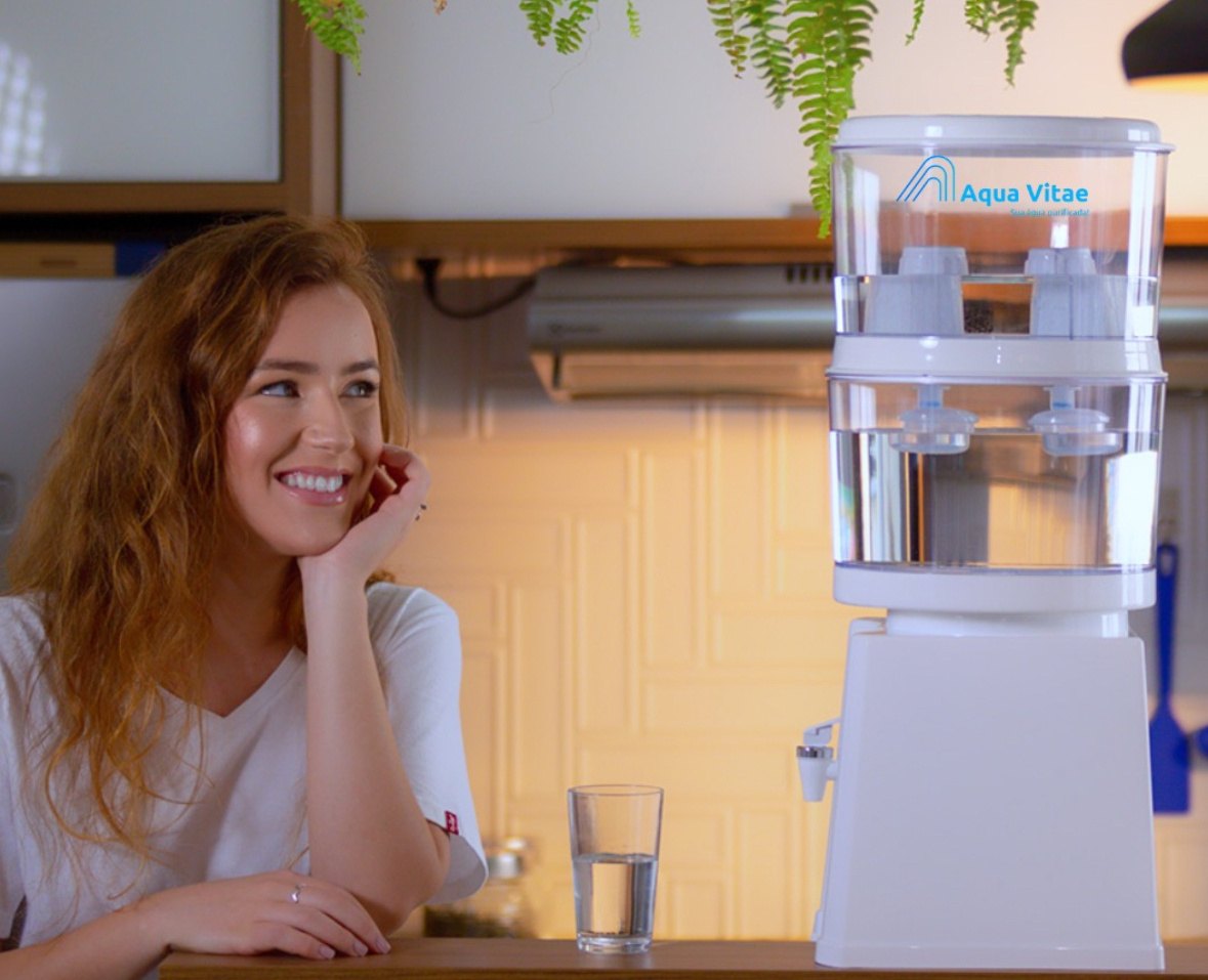 Mulher sorrindo na cozinha ao lado de um filtro Aqua Vitae e de um copo de água